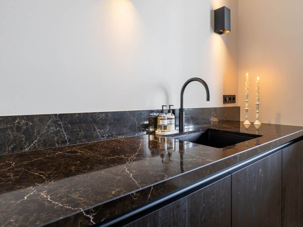 Impressie van een luxe aanrecht met een zwart marmeren werkblad en donker houten kastdeuren. Het aanrecht is ingericht met een design wasbak en een zwarte Quooker.