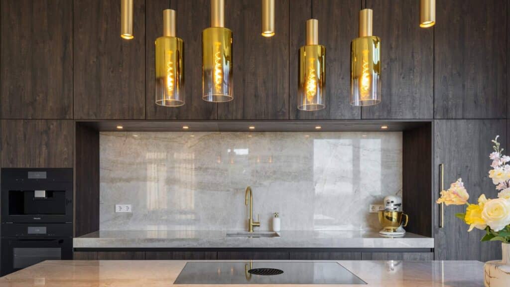 Luxe keuken met donker hout en glanzend marmer met gouden aders. We zien ook goudkleurige plafondlampen en een Quooker in goud.