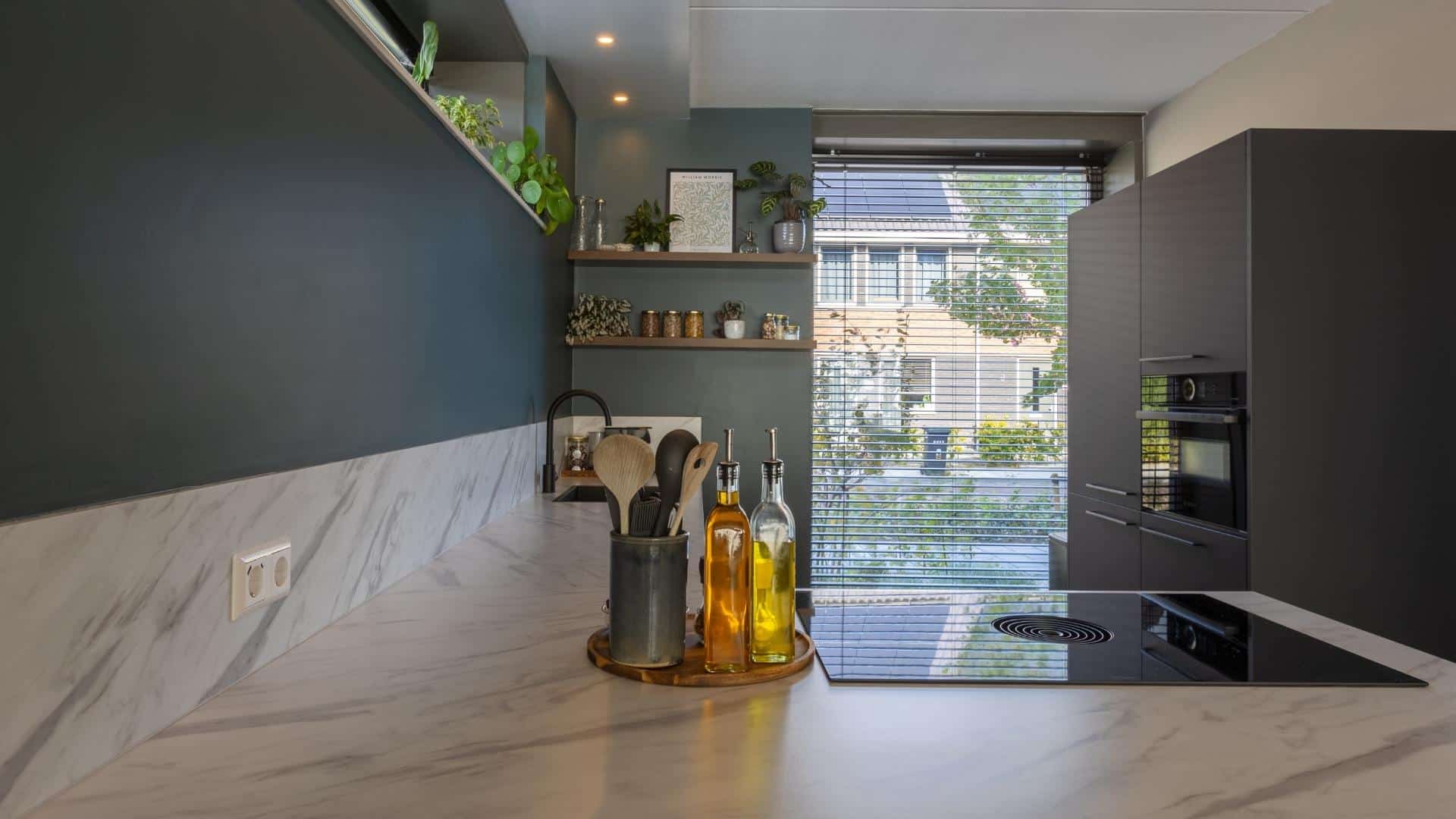 Impressie van een moderne hoekkeuken met natuursteenlook blad, kookplaat met werkbladafzuiging en zwarte kastenwand met inbouw apparatuur.