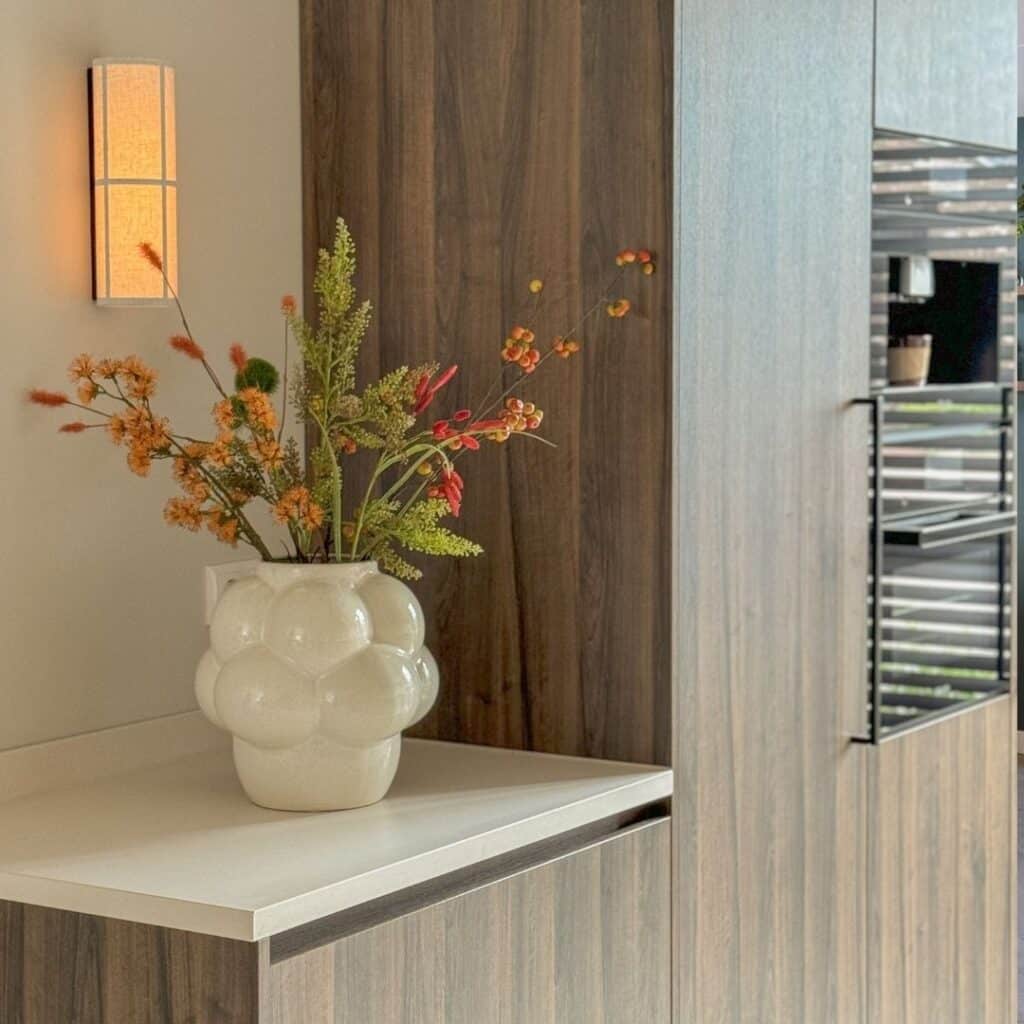 Impressie van een houten kastenwand in een Japandi keuken. De kastenwand heeft inbouw ovens, een koelkast en een lagere kast met een decoratieve vaas. Daarboven zorgt een wandlamp voor warme sfeerverlichting.