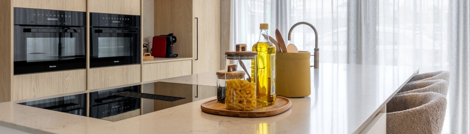 Impressie van een Japandi stijl keuken met kookeiland, natuursteenlook werkblad en licht eiken kastenwand met inbouw ovens.