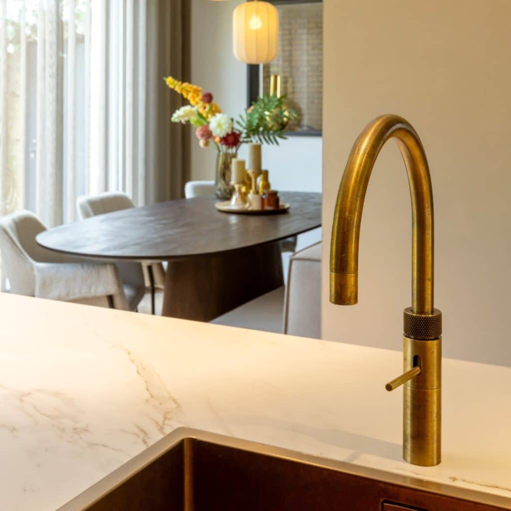 Impressie van een Quooker in messing patina finisch. Op de achtergrond zien we een eethoek met ovale houten tafel.