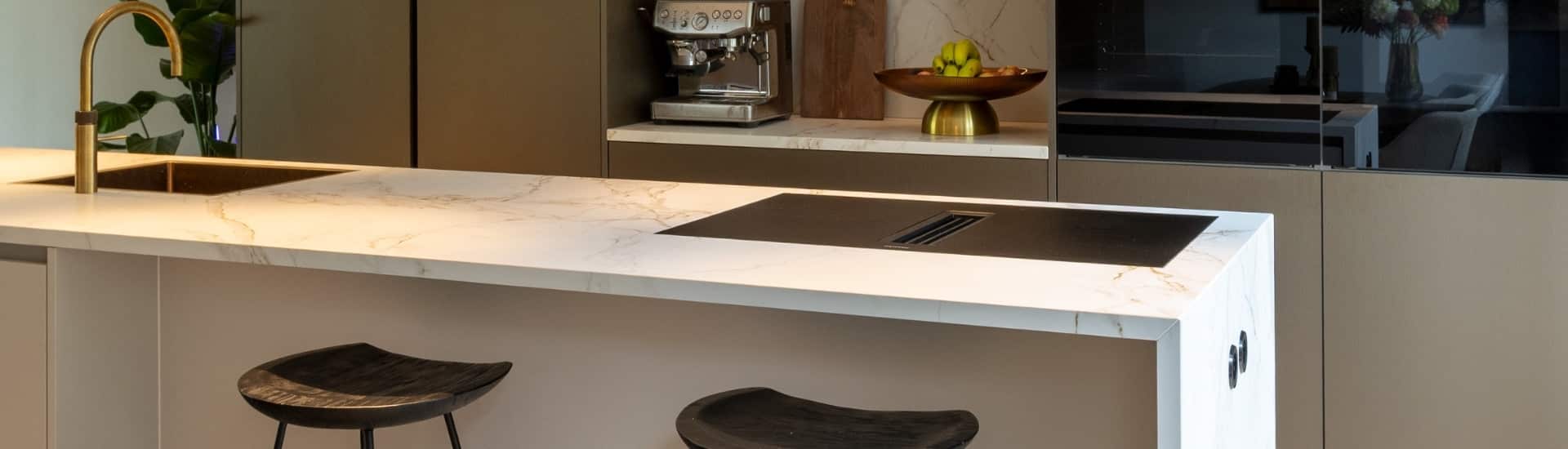 Impressie van een luxe keuken, voorzien van een kookeiland met bar. Het eiland heeft een marmerlook blad, met kookplaat en wasbak met Quooker in messing patina finish.