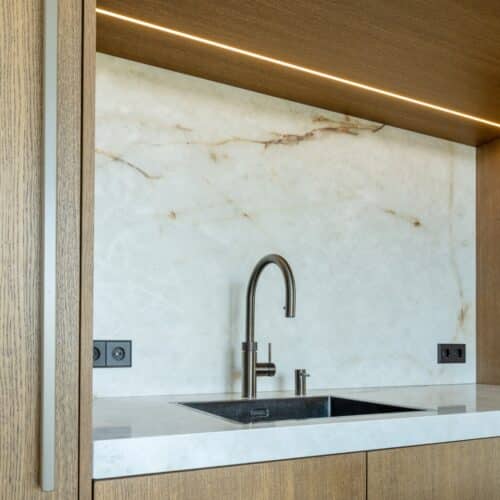 Impressie van een licht eiken kastenwand met nis, voorzien van marmerlook blad en achterwand, en ingericht met een design wasbak en gunmetal Quooker.