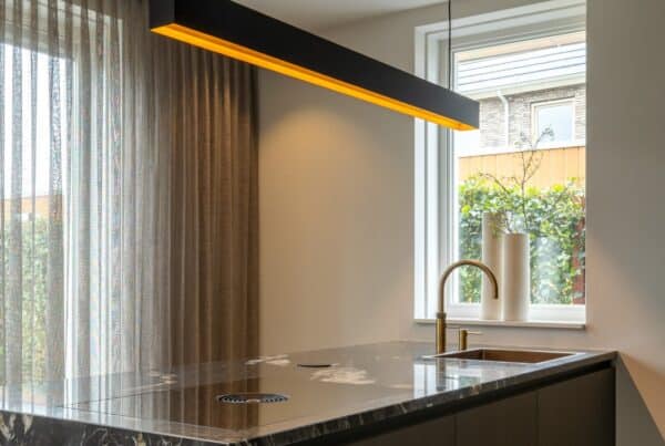 Impressie van een kookeiland met zwart marmerlook blad en zijwand, een Quooker met verweerde messing finish, een kookplaat met werkbladafzuiging en een zwarte designlamp boven het eiland met warm licht.