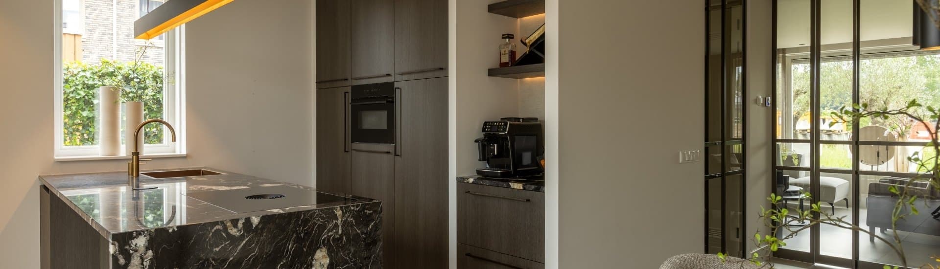 Impressie van een Hotel Chique keuken, voorzien van een kookeiland met zwart marmerlook blad en zijwand, een Quooker met verweerde messing finish, een kookplaat met werkbladafzuiging, een zwarte kastenwand met inbouwoven, een koffiecorner en een zwarte designlamp boven het eiland met warm licht.