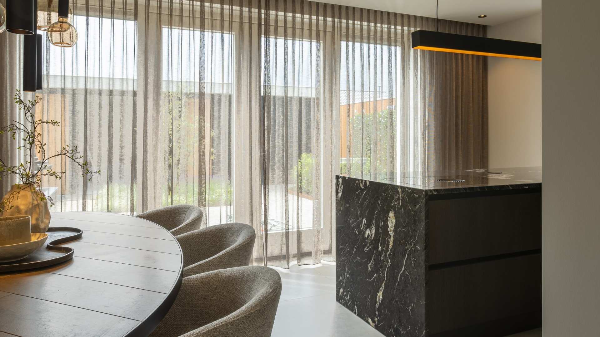 Moderne leefruimte, voorzien van zithoek met ovale houten tafel, met daarnaast een moderne keuken met zwart marmerlook kookeiland. Boven het eiland zien we een designlamp met natuurlijk licht.