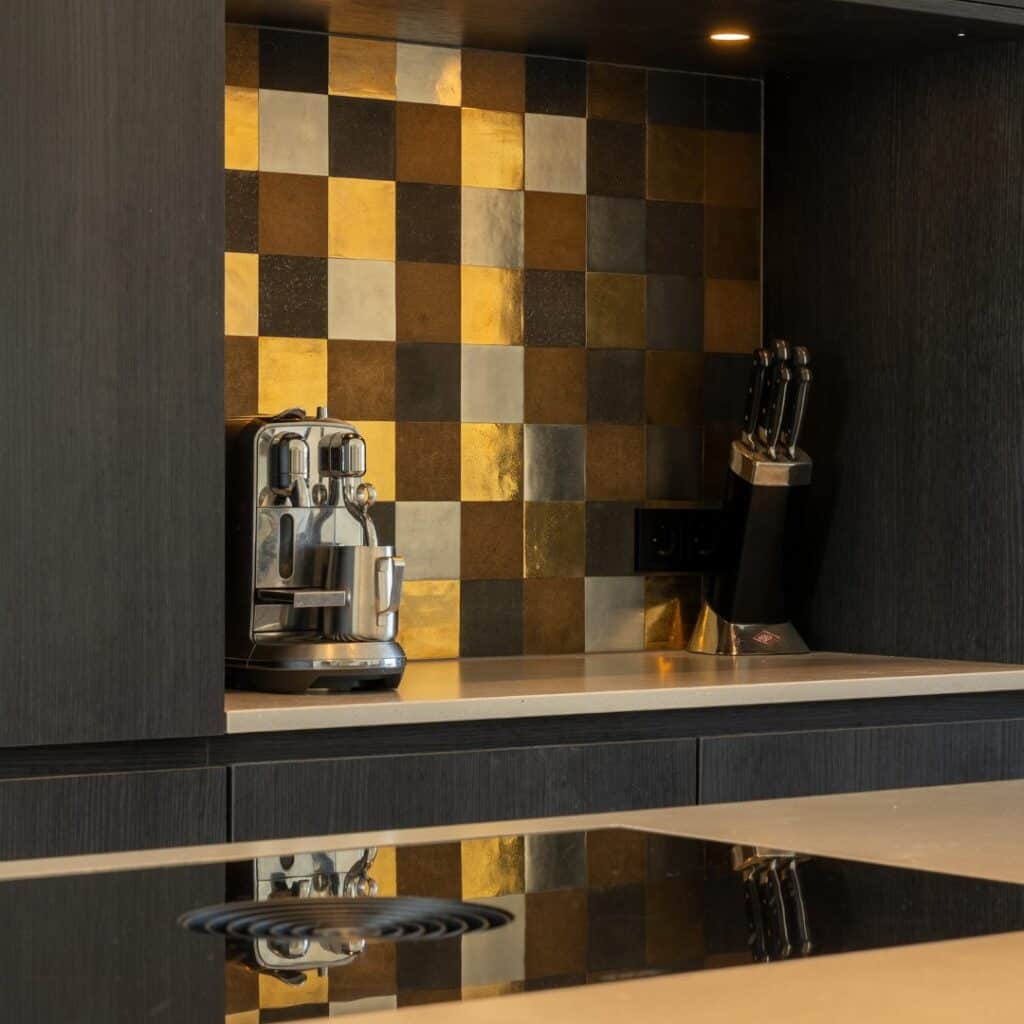 Impressie van een keuken nis in een zwarte kastenwand. De nis heeft een natuursteenlook werkblad en een achterwand met decoratieve tegeltjes, in de kleuren bruin, goud, zwart en grijs in een willekeurig patroon.
