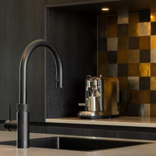 Impressie van een eiland met een zwarte Quooker, met op de achtergrond een keuken nis in een zwarte kastenwand. De nis heeft een natuursteenlook werkblad en een achterwand met decoratieve tegeltjes, in de kleuren bruin, goud, zwart en grijs in een willekeurig patroon.